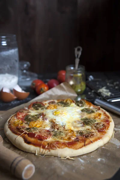 Fırın kağıdı üzerinde siyah ahşap bir masa üzerinde taze pişmiş pizza yatıyor. Oklavanın yanında. Arkasında domates, pesto ve un konserveleri ve yumurta kabukları var..