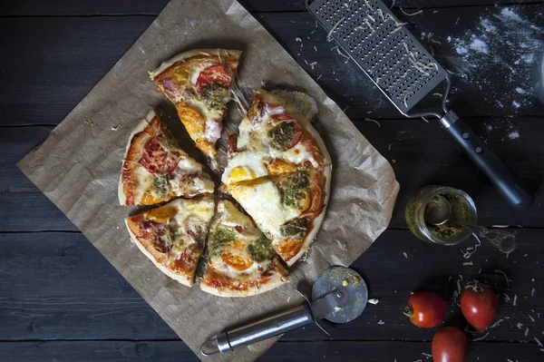 Siyah ahşap bir masa üzerinde bir fırın kağıdı üzerinde yatıyor dilimlenmiş taze pişmiş pizza, üst görünümü. Yakınlarda bir pizza bıçağı, bir rende domates ve bir kavanoz pesto var..