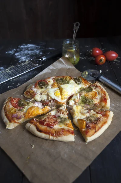 Dilimler halinde kesilmiş taze pişmiş pizza fırın kağıdı üzerinde siyah ahşap bir masa üzerinde yatıyor. Pizza bıçağının yanında. Domatesarkasında, pesto ve bir rende ile bir kavanoz.