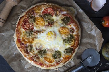 Siyah ahşap bir masa üzerinde pişirme kağıdı üzerinde yatan bir taze pişmiş pizza üst görünümü. Bir oklava, bir pizza bıçağı, yumurta kabukları, domates ve pesto bir kavanoz yakın yatırılır.