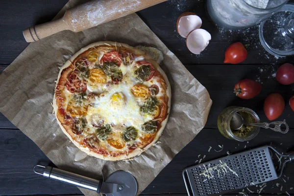 Siyah ahşap bir masa üzerinde pişirme kağıdı üzerinde yatan bir taze pişmiş pizza üst görünümü. Bir oklava, yumurta kabukları, domates, pesto ve un ve bir rende ile kavanoz yakınında.