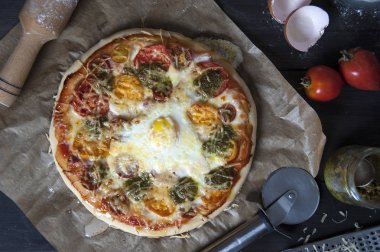 Siyah ahşap bir masa üzerinde pişirme kağıdı üzerinde yatan bir taze pişmiş pizza üst görünümü. Yakınlarda bir oklava, pizza bıçağı, yumurta kabuğu, domates ve bir kavanoz pesto yer alır..