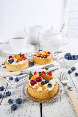 Krema ve taze çilek ile iştah açıcı hamur işleri bir kesme tahtası ve beyaz ahşap bir masa üzerinde gümüş bir tabak üzerinde yalan. Beyaz çay seti ve bir kase yaban mersini arkasında..