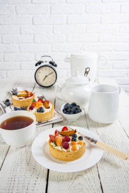 Beyaz natürmort. Krema ve taze çilek ile iştah açıcı kek bir marvelka ahşap bir masa ve bir kesme tahtası üzerinde yatıyor. Yakındaki çin servisi ve çalar saat. Tuğladan bir duvarın arkasında.