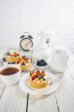 Beyaz natürmort. Krema ve taze çilek ile iştah açıcı kek bir marvelka ahşap bir masa ve bir kesme tahtası üzerinde yatıyor. Bir fincan çay, servis ve çalar saatin yanında. Tuğladan bir duvarın arkasında.