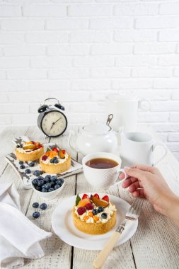 Beyaz natürmort. Bir tabak ve kesme tahtası ahşap bir masada çilek ile lezzetli hamur işleri vardır. Yakınlarda bir çay servisi ve pamuklu peçete bulunmaktadır. Bir kadın el fincan için uzanır.