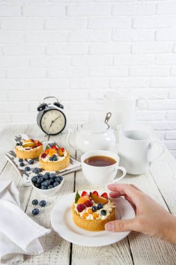Beyaz natürmort. Bir tabak ve kesme tahtası ahşap bir masada çilek ile lezzetli hamur işleri vardır. Çay setinin arkasında bir servis ve bir saat var. Bir kadın el pasta için uzanır.