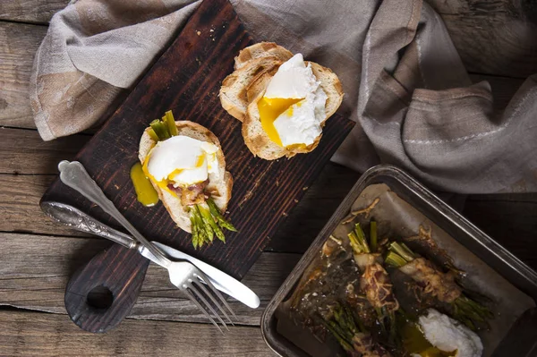 Top uitzicht op wit brood toast met gepocheerd ei en asperges gebakken met spek, liggend op een snijplank op de tafel. In de buurt van een vork met een mes, een linnen handdoek en een gerecht met asperges. — Stockfoto