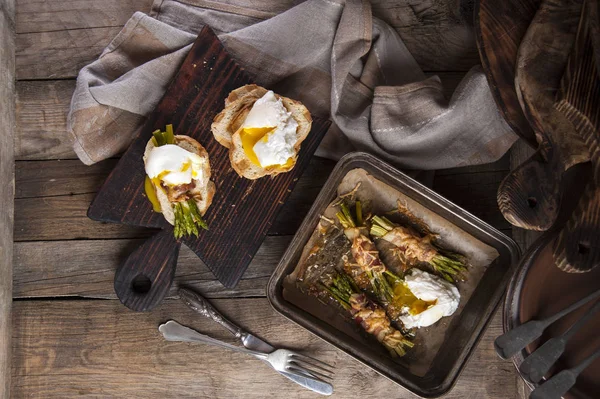 Draufsicht auf einen alten Holztisch, auf dem eine Auflaufform mit gebackenen Speckröllchen mit Spargel und pochiertem Ei steht. neben den Toasts mit diesen Zutaten auf einem Schneidebrett. — Stockfoto