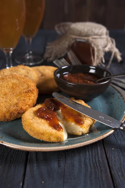 Een kom met gebakken Camembert en aardbei jam in een kom is op een oude houten tafel. In de buurt van het mes. Achter een potje jam, glazen bier. — Stockfoto