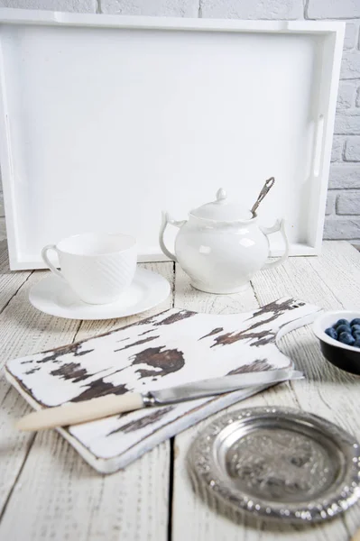 Naturaleza muerta en blanco: sobre una mesa de madera blanca hay un juego de té, un tazón con arándanos y un platillo de plata. En el centro hay una tabla de cortar con un cuchillo . — Foto de Stock