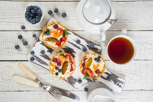 Beyaz ahşap bir masaüzerinde üst görünümü. Bir kesme tahtası üzerinde krem ve taze çilek ile shortcake hamur işleri vardır. Yakında beyaz çay servisi, yaban mersini ve çatal bıçak takımı bir kase.