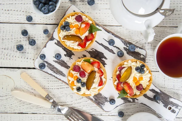 Beyaz ahşap bir masaüzerinde üst görünümü. Eski bir kesme tahtası üzerinde krem ve taze çilek ile shortcake hamur işleri vardır. Yakında beyaz çay servisi, yaban mersini ve çatal bıçak takımı bir kase.