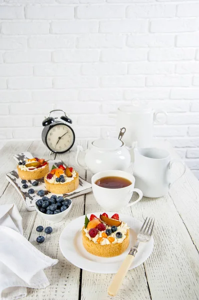 Beyaz natürmort. Bir tabak ve kesme tahtası ahşap bir masa üzerinde lezzetli hamur işleri vardır. Bir fincan çay, servis ve pamuklu peçetenin yanında. Tuğladan bir duvarın arkasında.