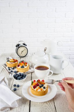 Beyaz natürmort. Bir tabak ve kesme tahtası ahşap bir masada çilek ile lezzetli hamur işleri vardır. Yakınlarda bir çay servisi ve pamuklu peçete bulunmaktadır. Çatallı kız pastayı kırıyor.