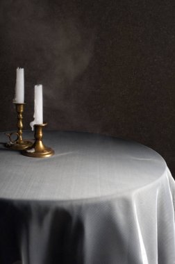 Beyaz bir masa örtüsü ile kaplı yuvarlak bir masa üzerine bir ışık ışını düşer. Üzerinde mumlu iki bronz şamdan var. Masanın yüzeyinden bir pus yükselir. Arka planda koyu kahverengi bir wal olduğunu