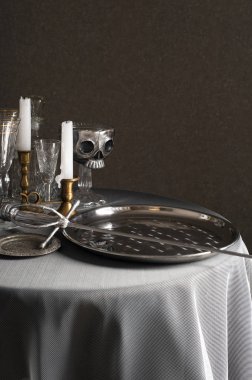 Beyaz bir masa örtüsü ile kaplı yuvarlak bir masa üzerinde gümüş tabak ve bir kılıç vardır. Yakınlarda mumlar, gözlükler ve bir stand üzerinde gümüş kafatası ile bronz şamdanlar vardır.
