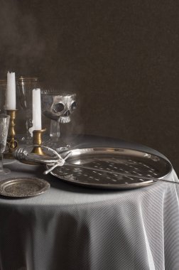 Beyaz bir masa örtüsü ile kaplı yuvarlak bir masa üzerinde gümüş tabak ve bir kılıç vardır. Yakınlarda mumlar, gözlükler ve bir stand üzerinde gümüş kafatası ile bronz şamdanlar vardır.