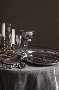 Beyaz bir masa örtüsü ile kaplı bir masa üzerinde gümüş tabak ve bir kılıç vardır. Yakında zincirli büyük bir metal madalyon vardır. Arkasında mumlar, gözlükler ve bir stand üzerinde gümüş kafatası ile mumluklar vardır.
