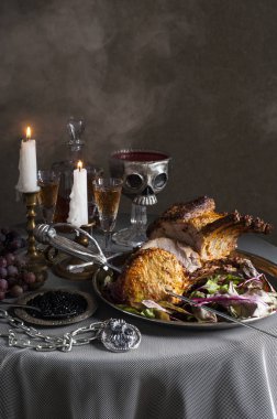 Bir yuvarlak üzerine, döşenmiş masa kuzu pişmiş raf ile bir tepsi. Yakınlarda şarap, mum ve üzüm kadehleri bulunmaktadır. Kasenin arkasında kafatası şeklinde bir kase kan var. Havada hafif bir pus var..