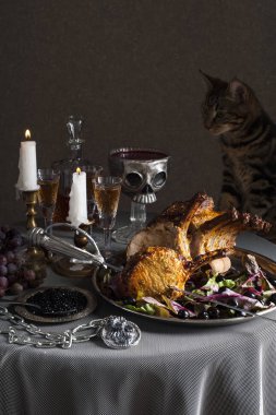 Bir kedi yuvarlak, masaya yatırılıyor. Ön planda bir kılıç ile delinmiş kuzu pişmiş raf ile bir tepsi. Arkasında şarap kadehleri, mumlar, üzümler ve kafatası şeklinde bir kase kan var..