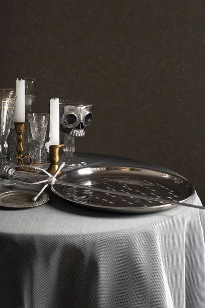 On a round table covered with a white tablecloth are silver plates and a sword. Nearby are bronze candlesticks with candles, glasses and a silver skull on a stand. — Stock Photo, Image