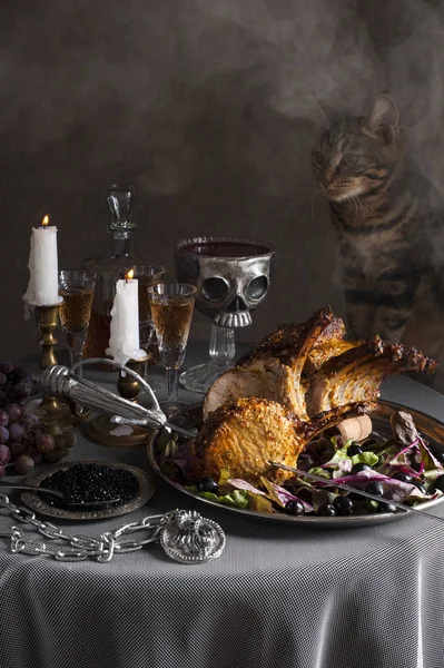 A cat sneaks onto a round table. In the foreground is a tray with a baked rack of lamb pierced with a saber. Behind are glasses of wine, candles and a skull-shaped bowl of blood. There is haze in the — Stock Photo, Image