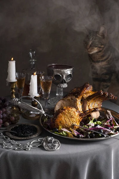 Een kat sluipt op een ronde tafel. Op de voorgrond is een dienblad met een gebakken lamsrack met een Saber doorboord. Achter zijn glazen wijn, kaarsen en een schedel vormige kom bloed. Er is nevel in de — Stockfoto