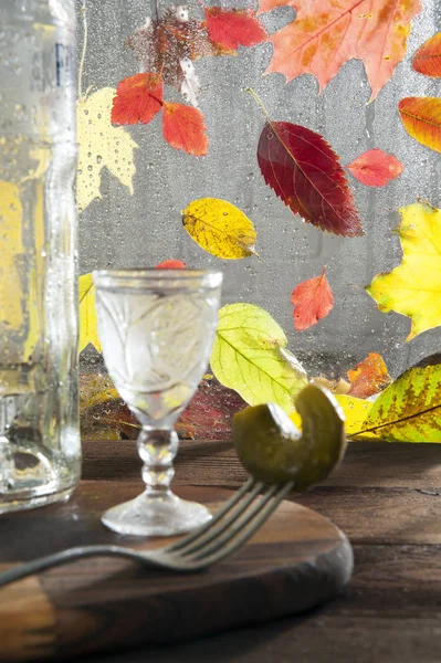 On a wooden table, lit by the sun, lies a cutting board on which stands a crystal glass with vodka. Nearby is a bottle and a fork with a chopped pickle. In the background, behind a wet window, leaf fa