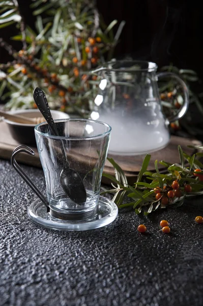 Auf einem schwarzen Tisch steht ein Teepaar aus Glas mit einem Löffel. In der Nähe steht eine mit Dampf gefüllte gläserne Teekanne auf einem hölzernen Schneidebrett. hinter einem Zweig Sanddorn, übersät mit reifen Beeren. — Stockfoto