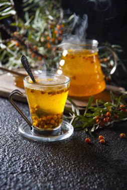 Siyah bir masada, sıcak deniz dikeni çayıyla dolu bir kesme tahtasında çaydanlık ve çaydanlık. Etrafında olgun çilek ile noktalı deniz dikeni dalları vardır. İçkiden buhar yükselir.