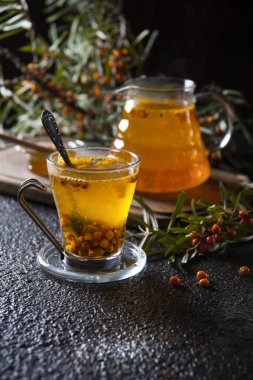 Siyah bir masada deniz dikeni çayı ile bir çay çifti. Bir bardak ta bir çay kaşığıdır. Çaydanlık bir çaydanlık kesme tahtasının üzerinde duruyor. Etrafında olgun çilek ile noktalı deniz dikeni dalları vardır.