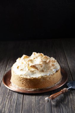 Kaba ahşap bir masanın üzerinde kremalı taze pişmiş bisküviyle birlikte bir tabak var. Pasta için spatulaya yakın. Arka planda siyah bir duvar var..