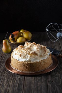 Sonbahar hala hayattır. Kaba ahşap bir masanın üzerinde taze pişmiş kremalı bisküviyle birlikte bakır bir tabak var. Pasta için spatulaya yakın. Arka planda olgun armutlar ve bir çırpı var..