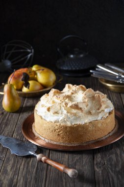 Koyu renk ahşap bir masada kremalı yeni pişmiş bir pastayla birlikte bakır bir tabak var. Pasta için spatulaya yakın. Arka planda olgun armutlar, bir çaydanlık, tabakta aletler ve çırpılmış bir çırpı var..