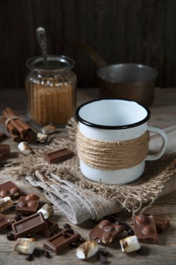 Ahşap bir masada, keten bir peçetenin üzerinde boş bir mineli kupa var. Etrafta çikolata küpleri, kahve taneleri ve kızarmış marşmelovlar var. Cam kavanozun arkasında şeker, kaşık, tarçın ve kepçeyle.