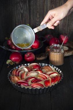 Elma dilimleriyle süslenmiş hamurla pişirme kabı ahşap bir masanın üzerinde duruyor. Arkadaki tabakta taze elma hasadı. Şekerli cam kavanozun yanında. Kız elinde erimiş tereyağlı bir kepçe tutuyor.