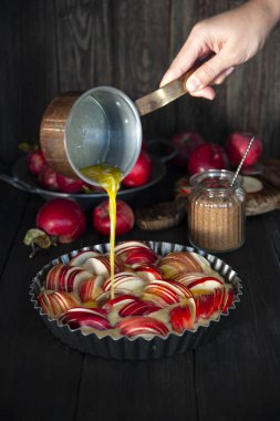 Elma dilimleriyle süslenmiş hamurla pişirme kabı ahşap bir masanın üzerinde duruyor. Arkadaki tabakta taze elma hasadı. Şekerli cam kavanozun yanında. Bir kız bir delikanlıdan erimiş tereyağı döker.