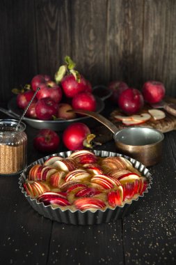Elma dilimleriyle süslenmiş hamurlu bir fırın tabağı, siyah ahşap bir masanın üzerinde duruyor. Arkadaki tabakta taze elma hasadı. Kesme tahtasının yanında, şekerli cam bir kavanoz ve bir kepçe..