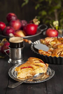 Bir dilim elmalı turta ve bir çay kaşığı siyah ahşap masadaki gümüş bir fincan tabağında yatıyor. Arka planda bir tabakta taze elmalar, bir bardak çay tutacağı ve bir tavada pasta ve bir yudum su var.