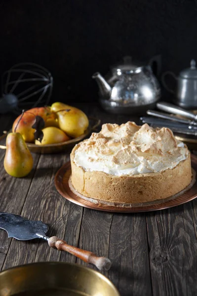 On a dark wood table is a copper dish with a biscuit cake with meringue cream. Near a spatula for cake and a plate. In the background are ripe pears, a teapot, cutlery in and a whisk.