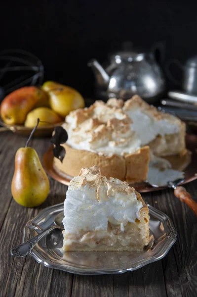 On a dark wood table there is a copper dish with a biscuit cake with meringue cream and a spatula for the cake. Before him is a sliced slice in a silver plate. Behind ripe pears, teapot and cutlery.