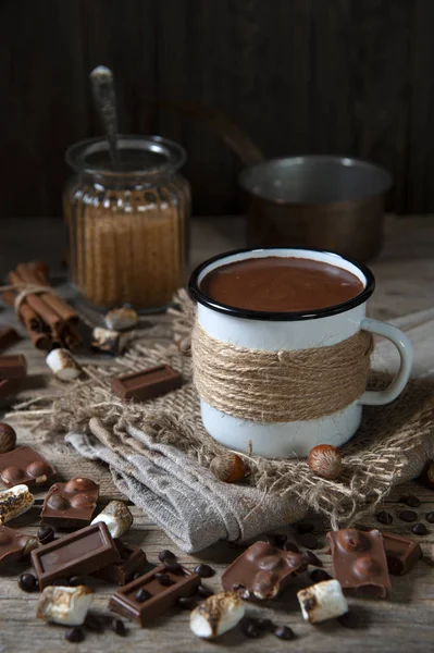 På bordet, på en servett, ligger en emaljerad mugg med varm choklad. Runt choklad, kaffekorn, rostade marshmallows och nötter. Bakom en glasburk med socker och en sked, kanel och en slev. — Stockfoto