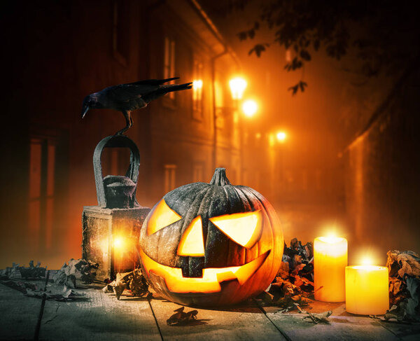 Scary horror background with halloween pumpkin jack o lantern, placed on wooden deck. Old town street on background with glowing lamps. Halloween spooky background.