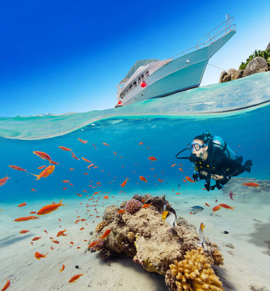 Under and above water surface view of woman diver. Underwater fauna and flora, marine life and exotic island with anchoring yacht on background