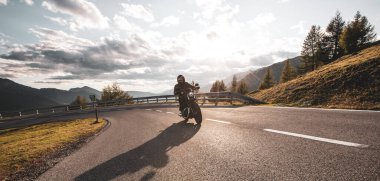Dağ yolunda motosiklet sürücüsü, geniş formatta. Dramatik gün batımı gökyüzü