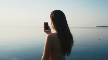 Tatile denize sıfır plaj üzerinde günbatımında onun Smartphone ile güzel genç kadın alarak resimler. Ağır çekim