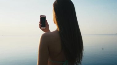Tatile denize sıfır plaj üzerinde günbatımında onun Smartphone ile güzel genç kadın alarak resimler. Ağır çekim