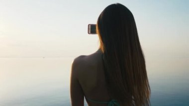 Tatile denize sıfır plaj üzerinde günbatımında onun Smartphone ile güzel genç kadın alarak resimler. Ağır çekim