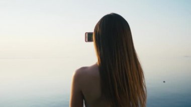 Tatile denize sıfır plaj üzerinde günbatımında onun Smartphone ile güzel genç kadın alarak resimler. Ağır çekim